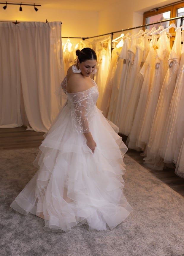 Entdecken Sie unser extravagantes Volantkleid mit eleganter Korsage und feinem Spitzentop. Die Rückenschnürung sorgt für den perfekten Halt und eine wunderschöne Silhouette. Brautmodengeschäft, Brautmode, Brautkleid, Brautkleider